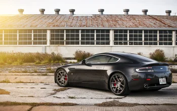 A dark Aston Martin Vantage against the backdrop of an abandoned hangar, bathed in the rays of the setting sun