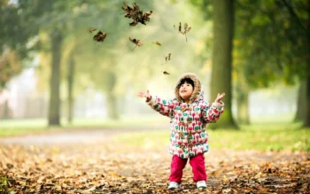 A little girl in a bright jacket is enjoying the falling autumn leaves in the park