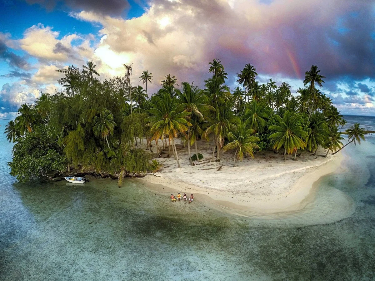 A group of people on the shore of a tropical atoll under a rainbow-colored sky