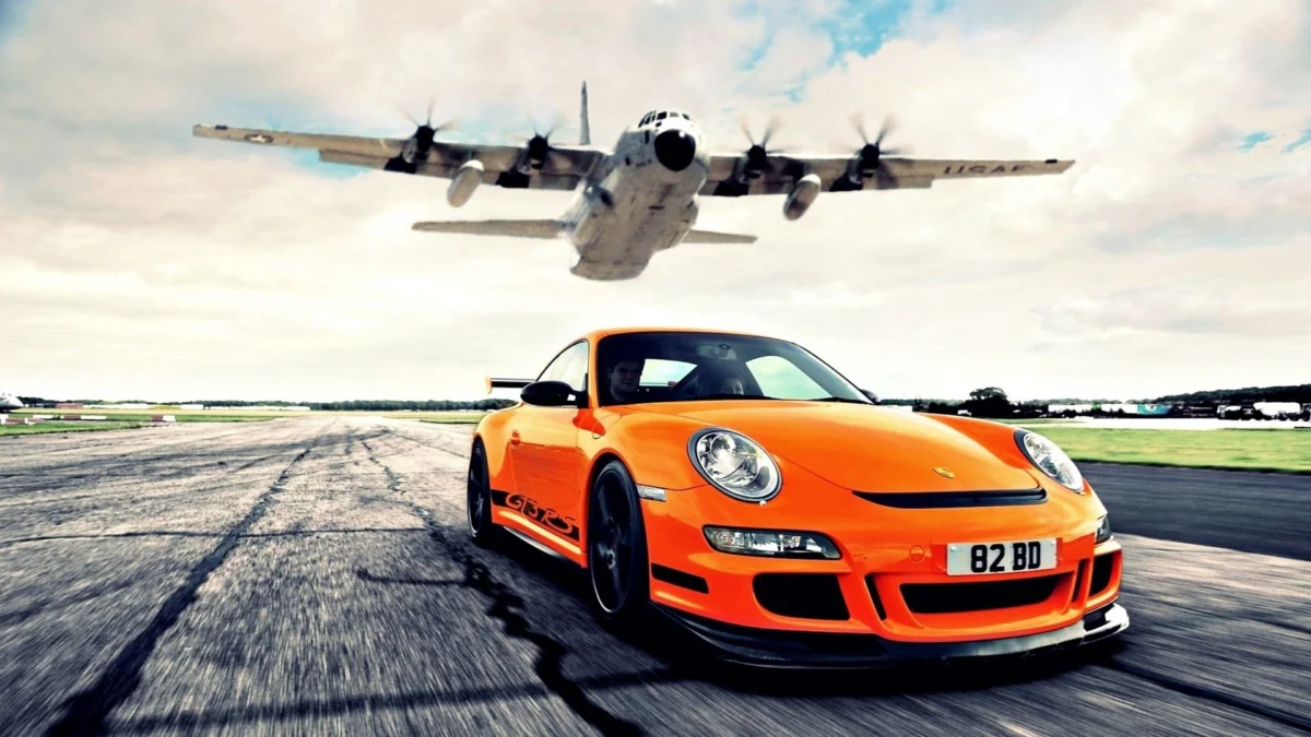 An orange Porsche 911 GT3 RS on the runway beneath a passing airplane