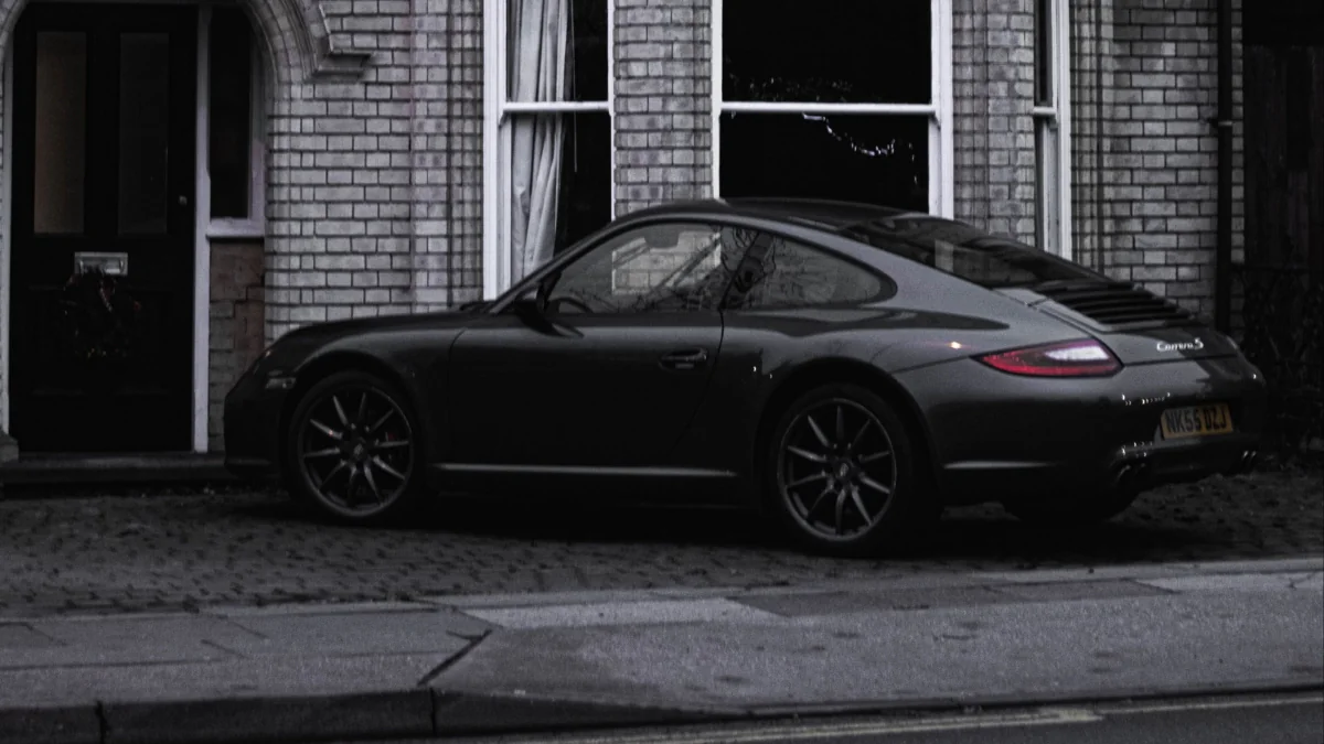 A dark-colored Porsche 911 Carrera S is parked next to a brick house