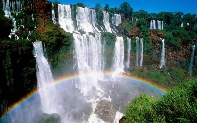 a rainbow over the powerful cascades of iguazu falls, iguazu, brazil, argentina, cliffs, herb, herbs, lawns, landscapes, rainbows, splashes, stones, waterfalls, nature